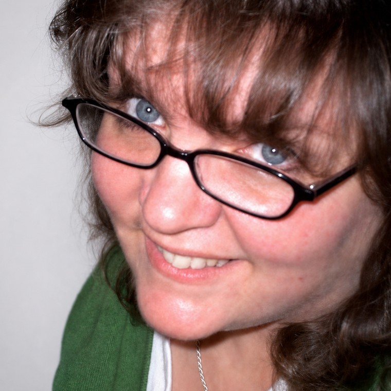 Kim Sponaugle — a smiling woman with dark curly hair and black-framed glasses, wearing a green cardigan